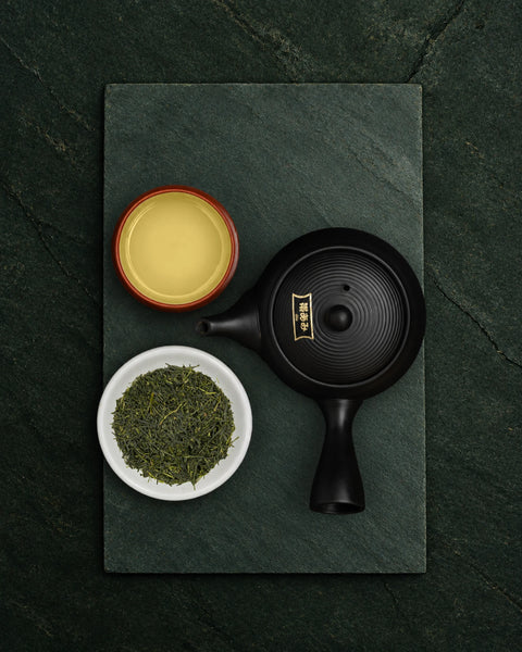 A black ceramic teapot with a gold emblem, a small white bowl of shincha loose green tea leaves, and a small red-rimmed cup of pale yellow tea, arranged on a dark green slate board against a dark green textured background.