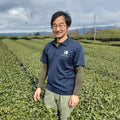 Miyazaki Farmerwearing a navy blue long-sleeve polo shirt with a white logo on the left chest stands in a lush green tea plantation. The shirt features the text 'Tea Master Club' and a stylized 'TM' logo.