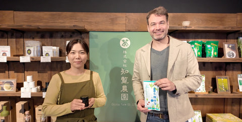 Nio Teas CEO and Nio Teas farmer stand in front of a wooden display wall with tea products. The woman wears a beige top and green apron, holding a small black cup.